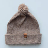 Light brown knitted baby hat with a fluffy pompom and a folded edge, handmade from 100% baby alpaca wool, featuring a soft vegan leather label.