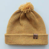 Handmade light yellow baby hat in soft 100% baby alpaca wool with a pompom, foldable edge, and brown vegan leather label, offering cozy warmth and cute design.