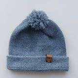 Handmade light blue baby hat in soft 100% baby alpaca wool with a pompom, foldable edge, and brown vegan leather label, offering warmth and comfort.