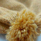 Close-up of the handmade light yellow baby hat’s pompom, made from soft 100% baby alpaca wool, with a foldable edge and brown vegan leather label.