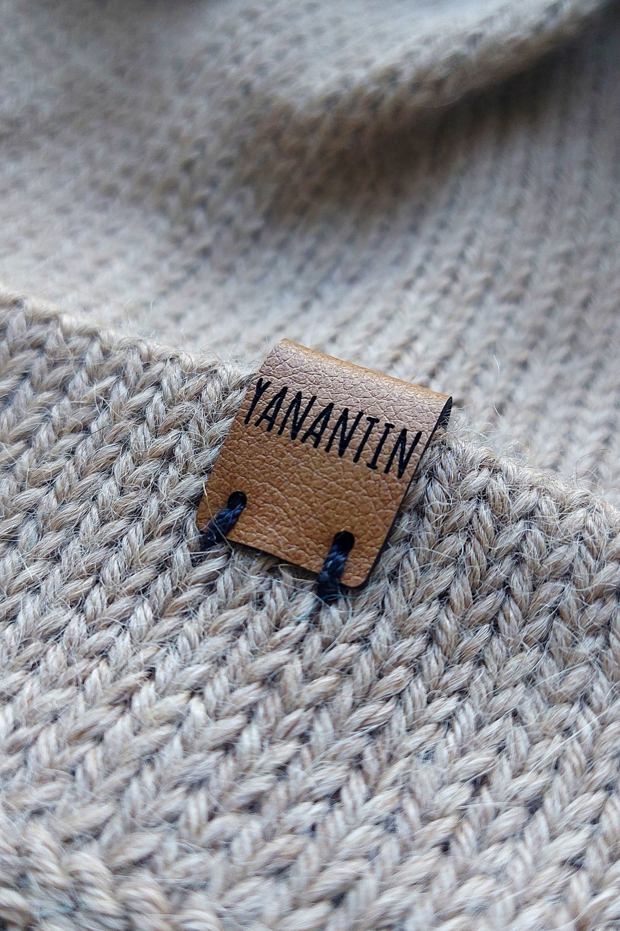 Close-up of the brown vegan leather label on a light brown knitted baby hat, made from 100% baby alpaca wool.