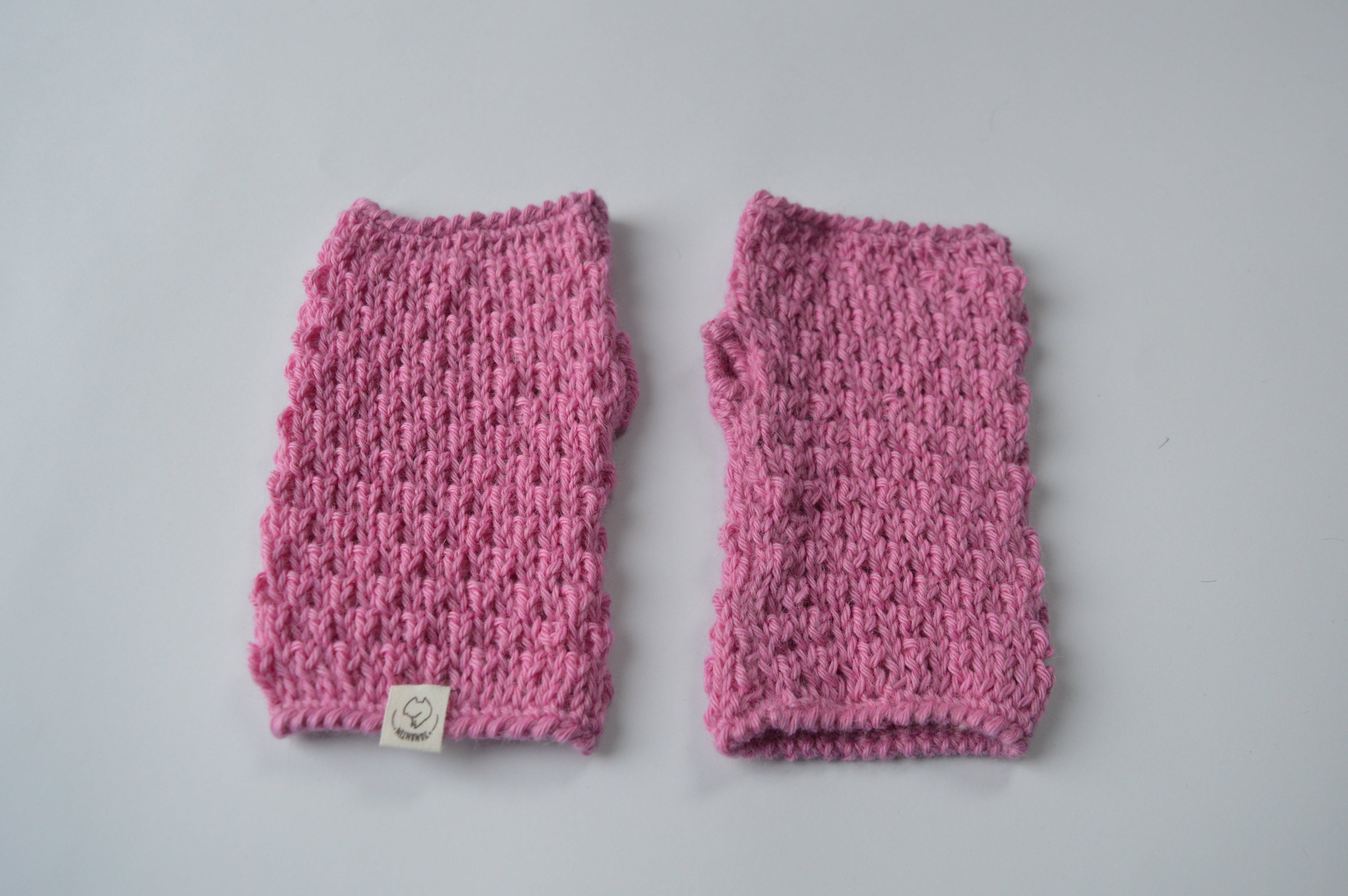 Pair of pink knitted fingerless mittens on a light background with an organic cotton label