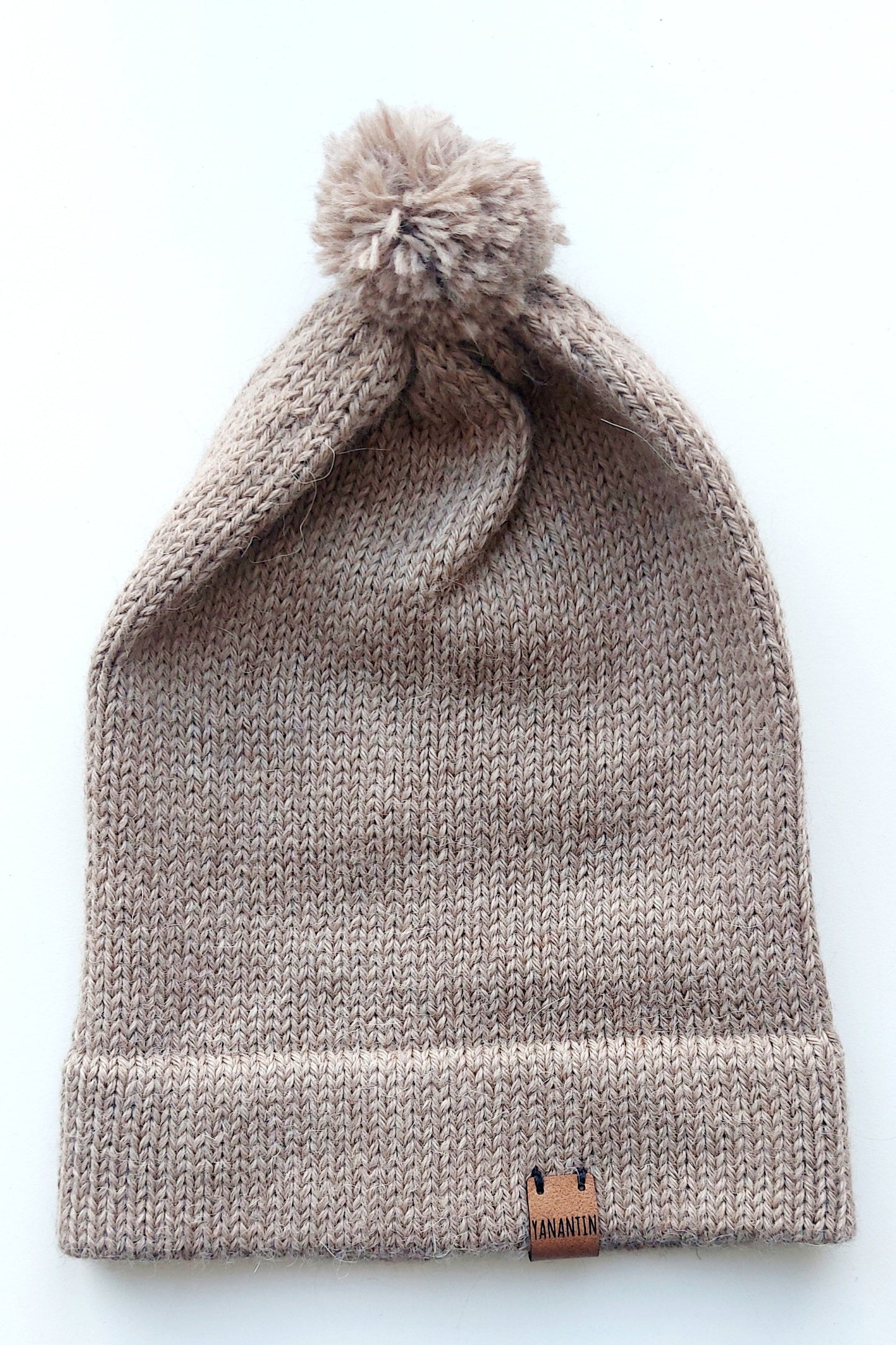 Light brown knitted baby hat with a fluffy pompom and a folded edge, handmade from 100% baby alpaca wool, featuring a soft vegan leather label.