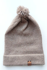 Light brown knitted baby hat with a fluffy pompom and a folded edge, handmade from 100% baby alpaca wool, featuring a soft vegan leather label.