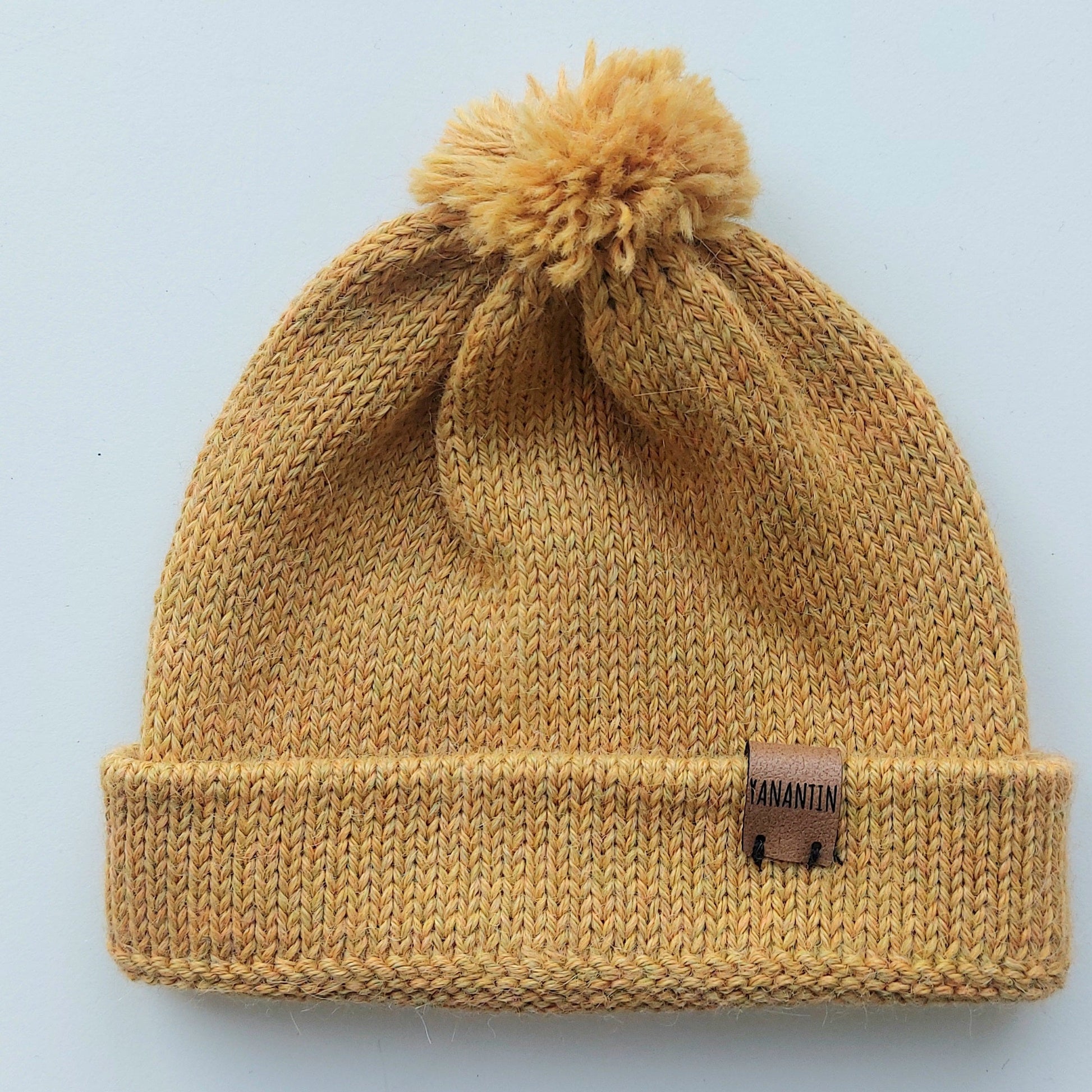 Handmade light yellow baby hat in soft 100% baby alpaca wool with a pompom, foldable edge, and brown vegan leather label, offering cozy warmth and cute design.

