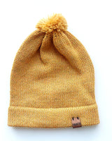 Handmade light yellow baby hat in soft 100% baby alpaca wool with a pompom, foldable edge, and brown vegan leather label, offering cozy warmth and cute design.
