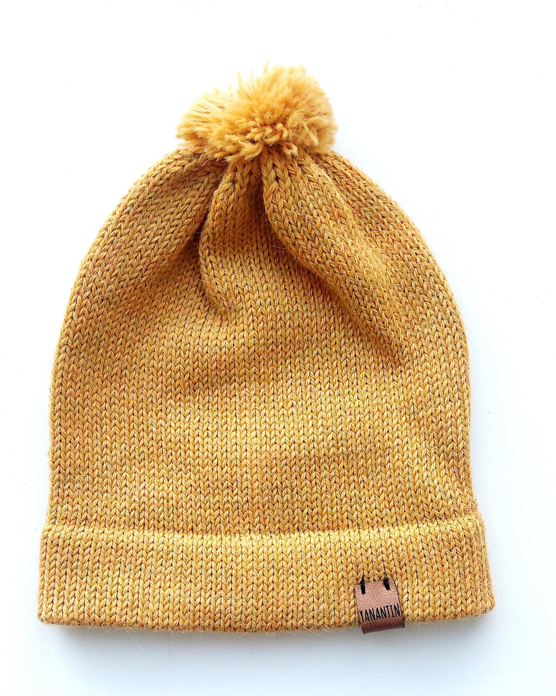 Handmade light yellow baby hat in soft 100% baby alpaca wool with a pompom, foldable edge, and brown vegan leather label, offering cozy warmth and cute design.
