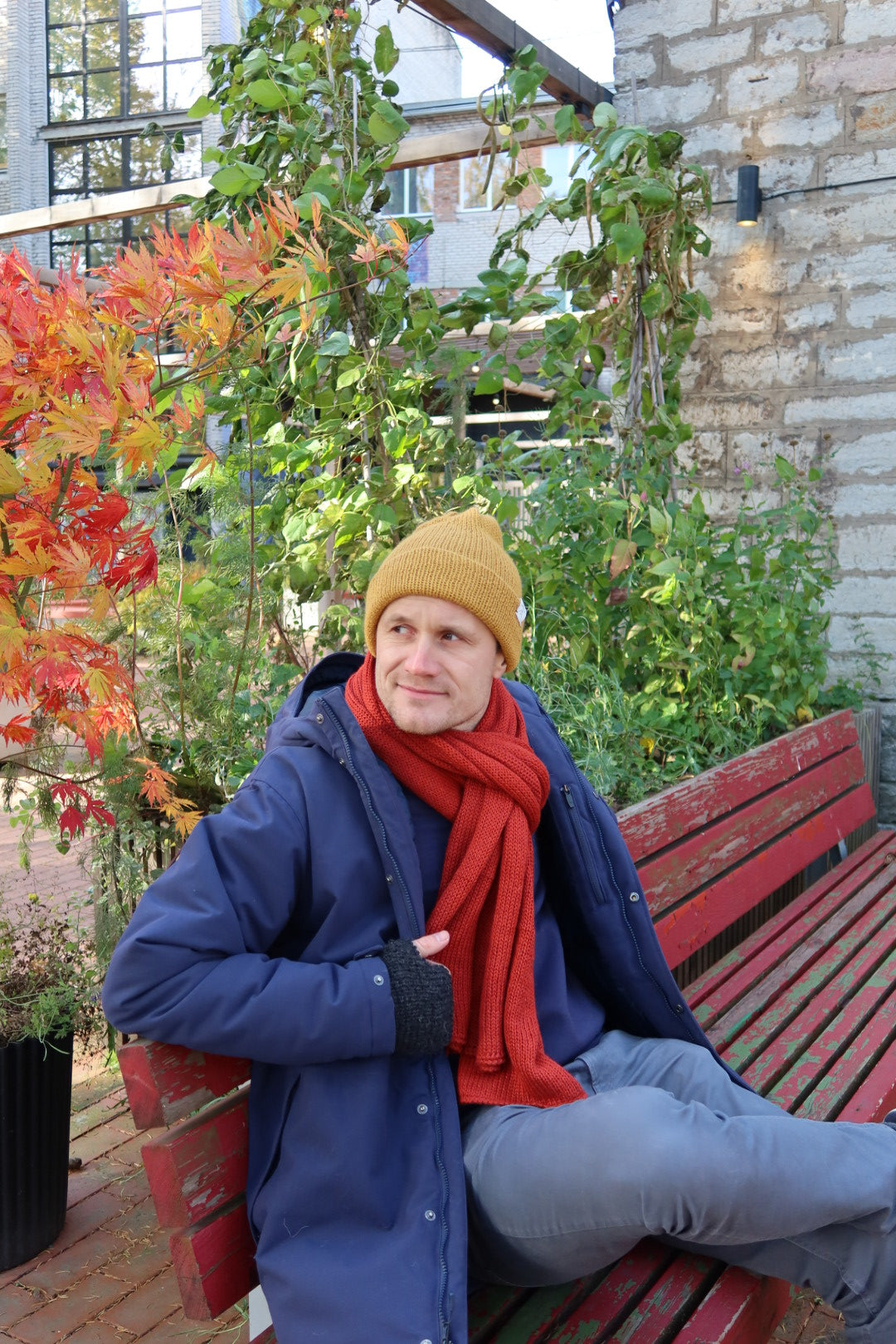 Young male wearing an yellow knitted hat and a yellow scarf sitting on a bench.