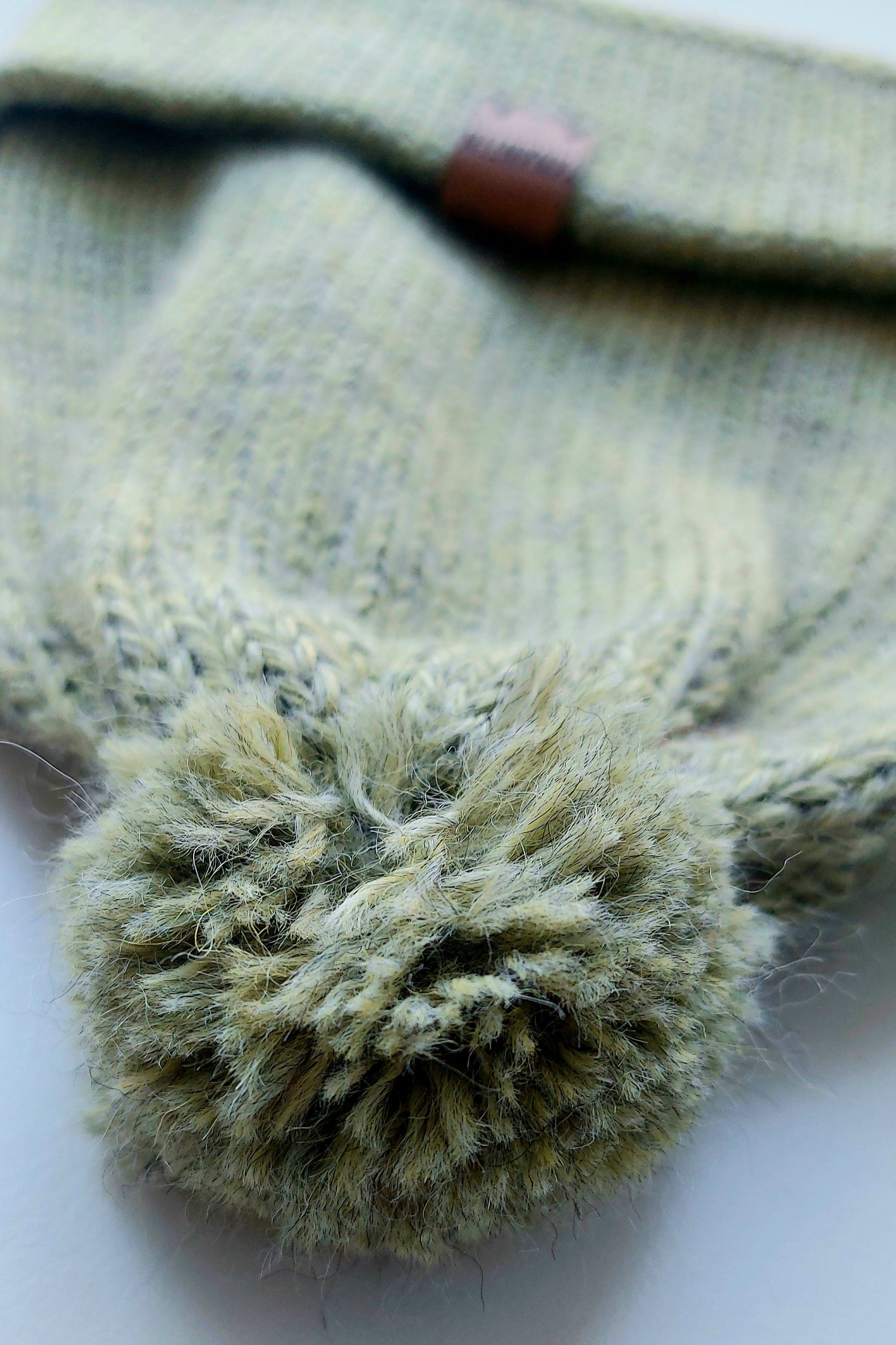 Close-up of the pompom on the handmade light green baby hat, made from soft 100% baby alpaca wool with a foldable edge and brown vegan leather label.