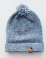 Handmade light blue baby hat in soft 100% baby alpaca wool with a pompom, foldable edge, and brown vegan leather label, offering warmth and comfort.