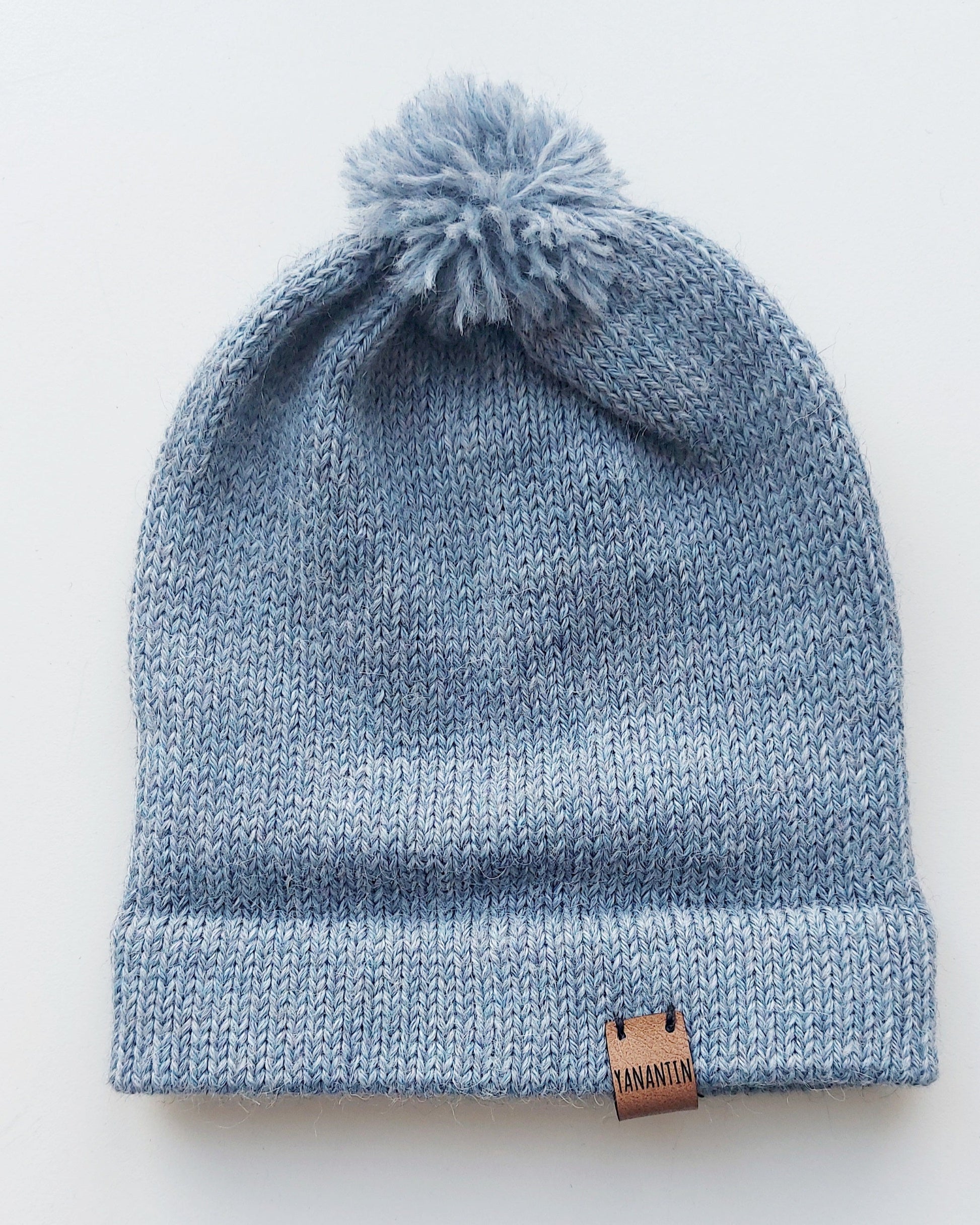 Handmade light blue baby hat in soft 100% baby alpaca wool with a pompom, foldable edge, and brown vegan leather label, offering warmth and comfort.