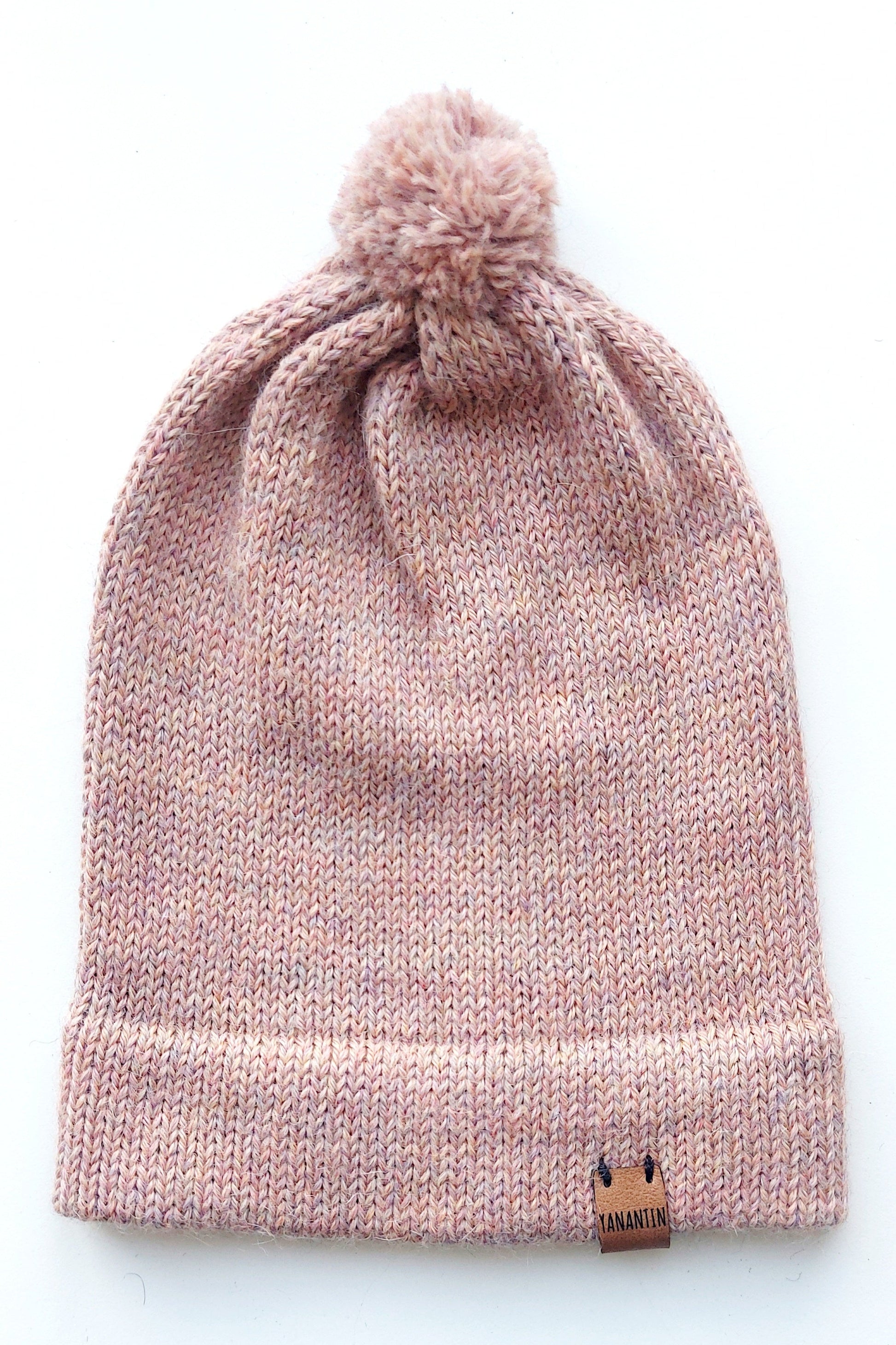Handmade light pink baby hat in soft 100% baby alpaca wool with a pompom, foldable edge, and brown vegan leather label, offering warmth and comfort.