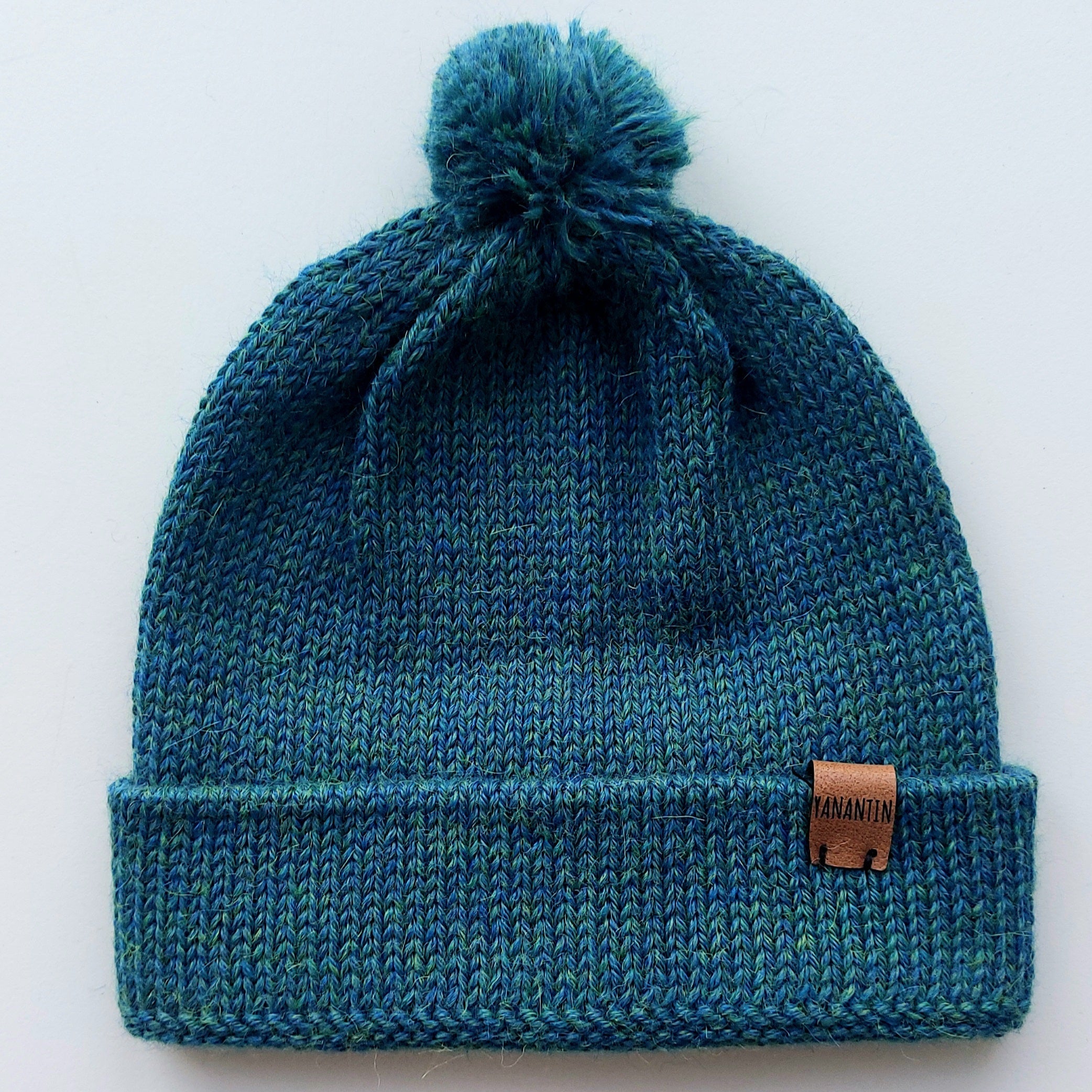 Handmade peacock blue baby hat in soft 100% baby alpaca wool with a pompom, foldable edge, and brown vegan leather label, offering warmth and comfort.