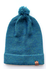 Handmade peacock blue baby hat in soft 100% baby alpaca wool with a pompom, foldable edge, and brown vegan leather label, offering warmth and comfort.